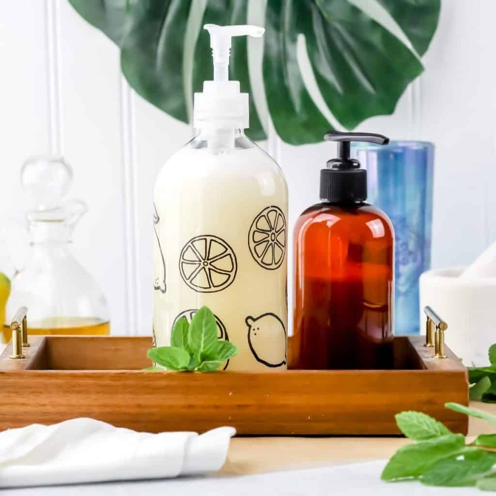 homemade hypoallergenic hand soap in a clear glass bottle is next to other bathroom bottles with a white towel and fresh mint leaves scattered around