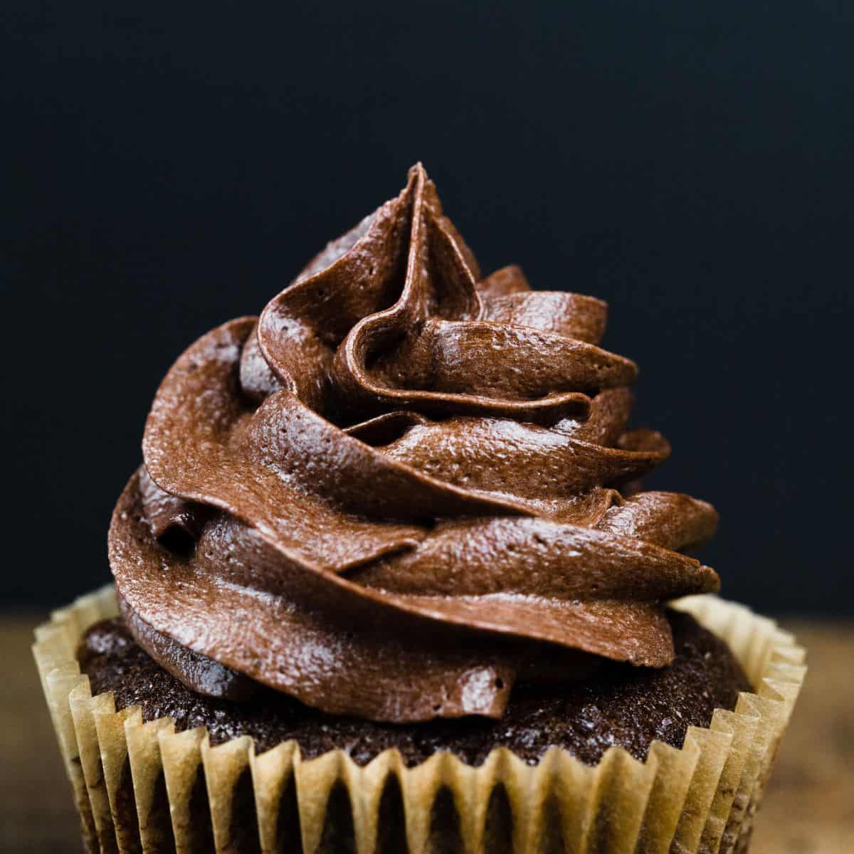 close up of a swirl of chocolate frosting on a chocolate cupcake in front of a black background so it looks like the frosting is in the spotlight