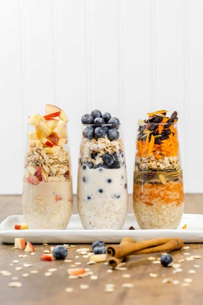 3 tall glasses filled with 3 flavors of overnight oats surrounded by ingredients like oats, cinnamon sticks, and blueberries