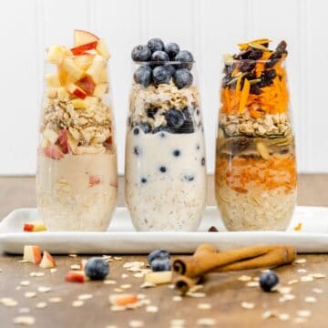 3 overnight oat flavors in tall skinny glasses on a plate on the kitchen table surrounded by ingredients like oats, cinnamon, and blueberries