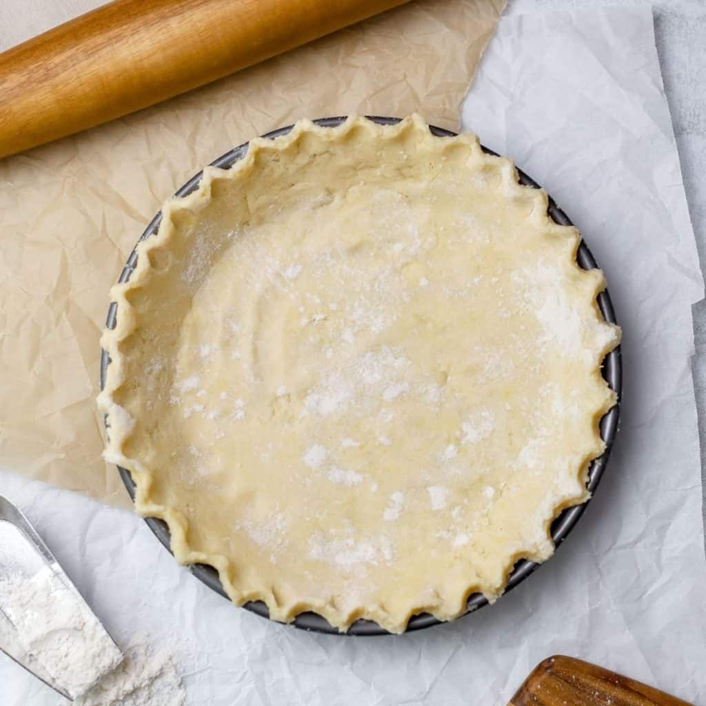 a pie crust is shown in a metal pan from the top down so you can see the simple crimping of the edges. items like a rolling pin and more flour surround the pie crust