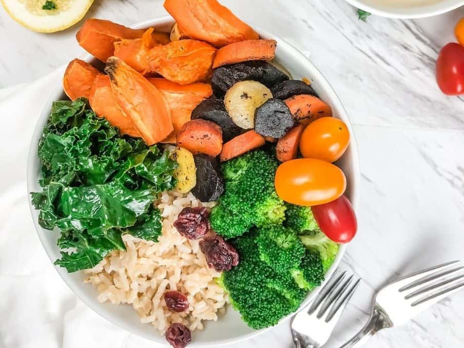 close up of rainbow roasted veggie bowl on a white countertop with forks and ingredients surround the bowl