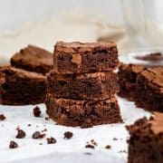 a stack of 3 gluten free brownies are in front of other brownies on white parchment paper on the kitchen countertop. chocolate chips and crumbs surround the brownies.