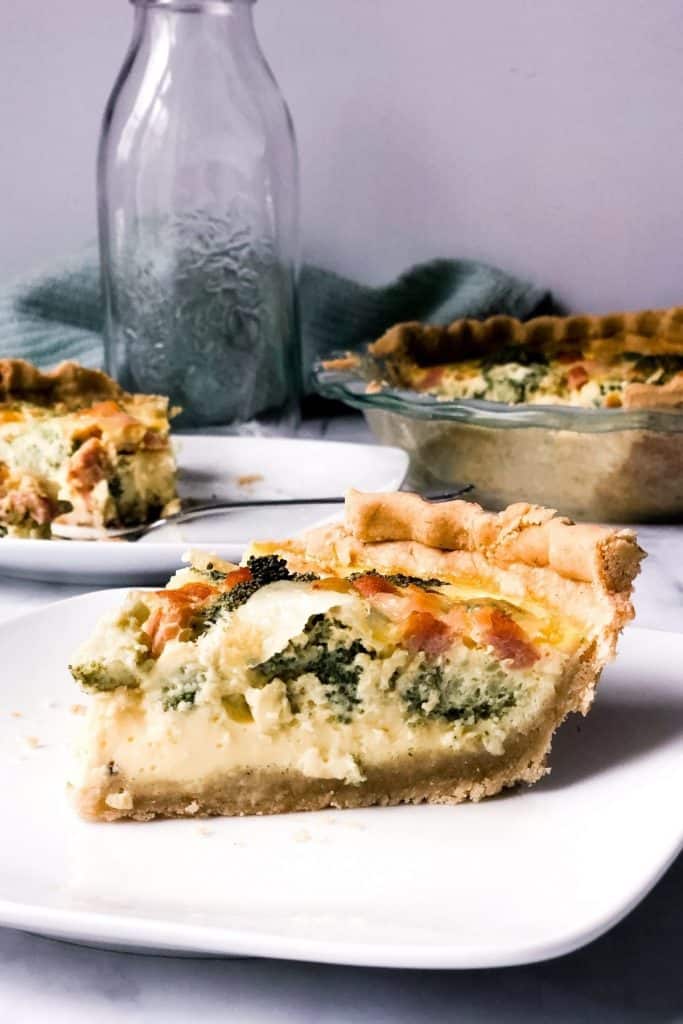 a slice of gluten free quiche rests on a white plate with more slices and the quiche blurred in the background