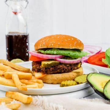 vegan black bean burger on a bun with many toppings is on a white plate with French fries and a glass of soda