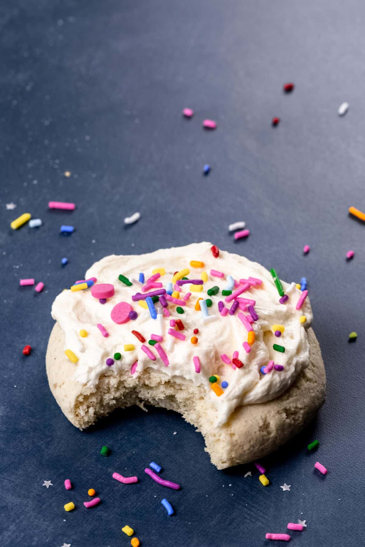 single gluten free soft sugar cookie with a bite taken out of it on a dark background surrounded by extra sprinkles