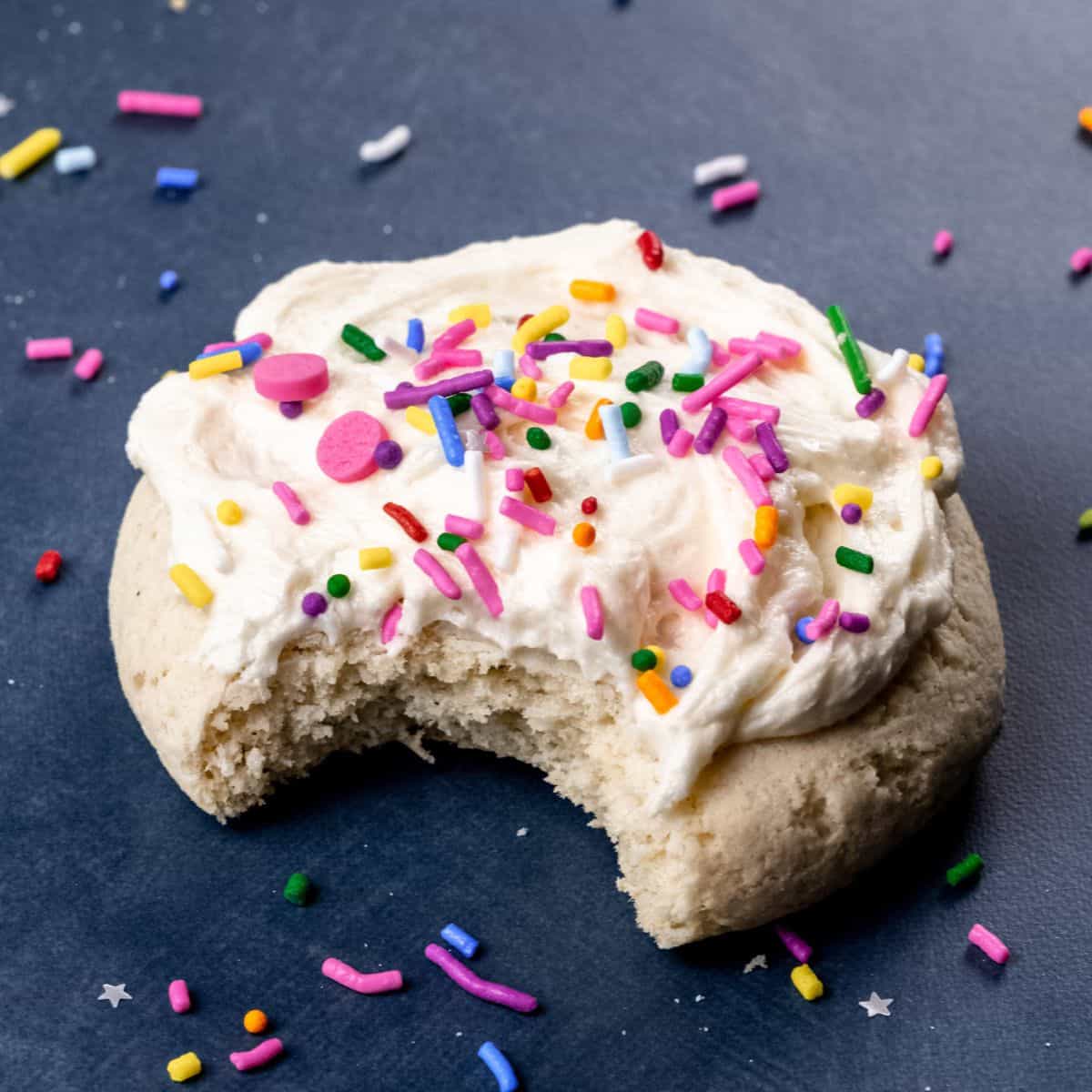 single gluten free soft sugar cookie on a dark background surrounded by sprinkles with a bite taken out of the cookie