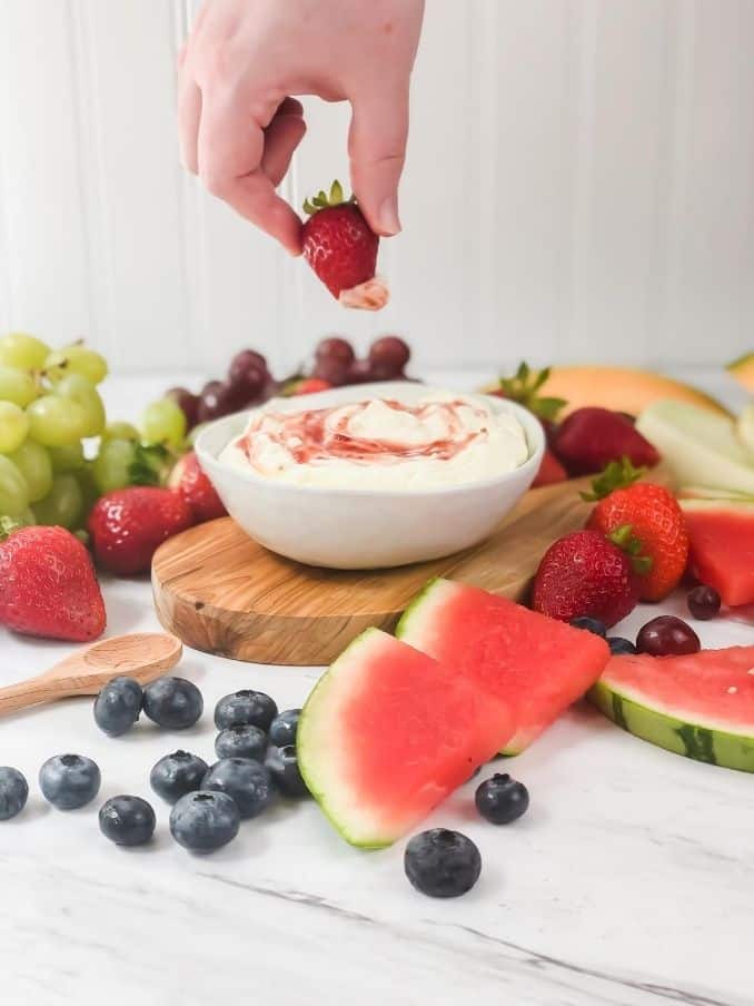 hand dipping a strawberry into 3 ingredient frui dip on table surrounded by fruit // livingbeyondallergies.com