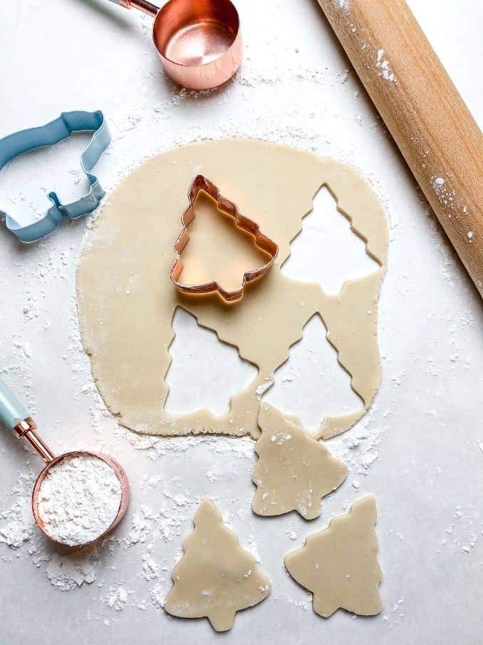 rolling out gluten free sugar cookies with tree shapes on the countertop surrounded by baking essentials // livingbeyondallergies.com