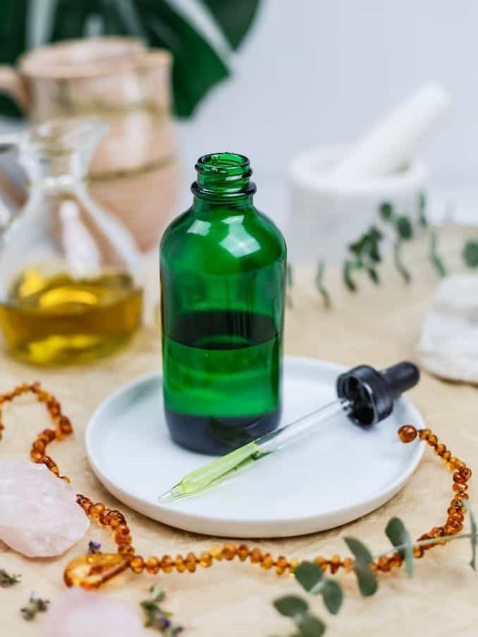 green bottle of body oil with the dropper rest on a round white plate with other oils and various bath items in the background