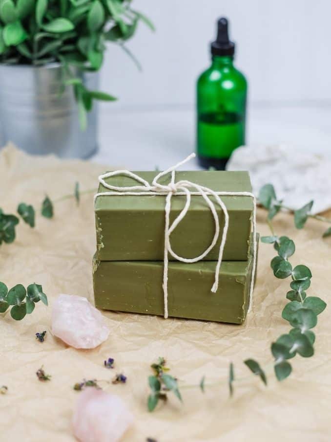 olive oil soap bars stacked on top of each other and wrapped in string surrounded by bath items on a crinkled brown parchment paper 