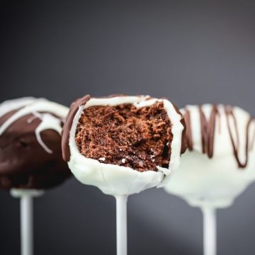3 gluten free chocolate cake pops are shown up close on sticks in front of a black background. the middle cake pop has a bite taken out of it to show off the chocolate cake on the inside