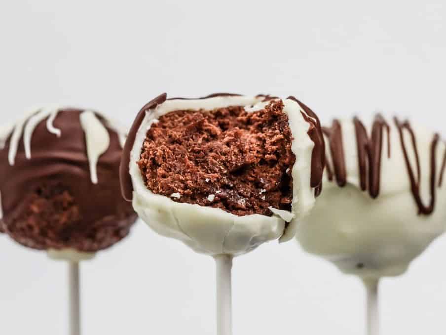 3 gluten free chocolate cake pops are shown in front of a white background and the front middle cake pop has a bite taken out of it so you can see the chocolate cake on the inside