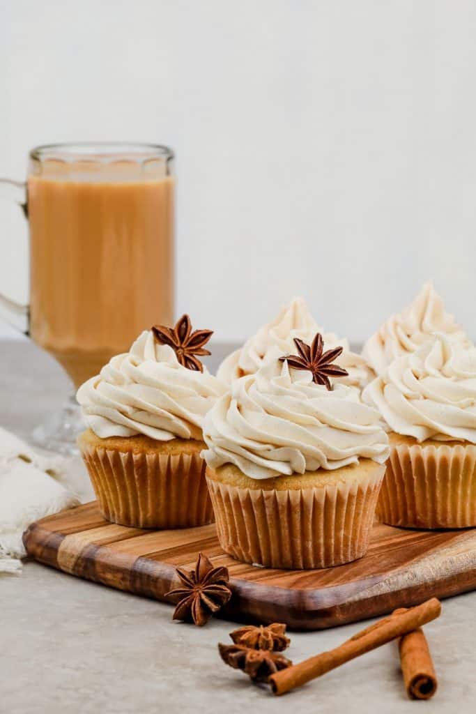 chai cupcakes rest on a wood board with a glass of chai in the background and cinnamon sticks and star anise pods around them
