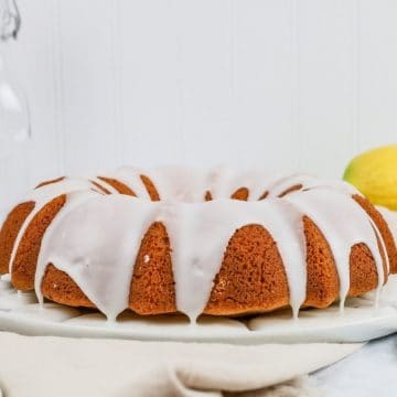 a gluten free lemon poppyseed cake tin a bundt shape is resting on a white plate on the kitchen countertop surrounded by a linen towel and lemons