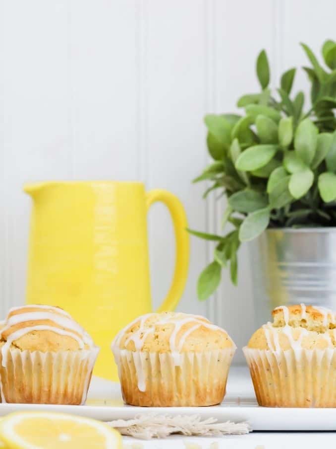 3 gluten free lemon poppyseed muffins are on a long white plate with lemon slices in front of them and a yellow kitchen and bright green plant in the background