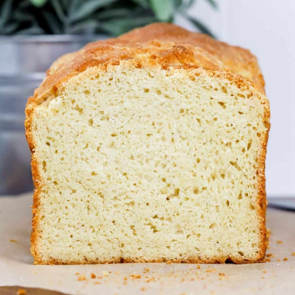 a loaf of gluten free bread is sliced open so you can see the perfect texture of the bread on the inside. It rests on brown parchment paper on the kitchen countertop in front of a green plant in a silver vase.