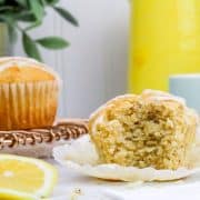 a gluten free lemon poppyseed muffin is shown with a bite taken out of it so you can see the inside texture of this muffin. More muffins and a yellow pitcher are in the background while lemon slices are seen in the foreground