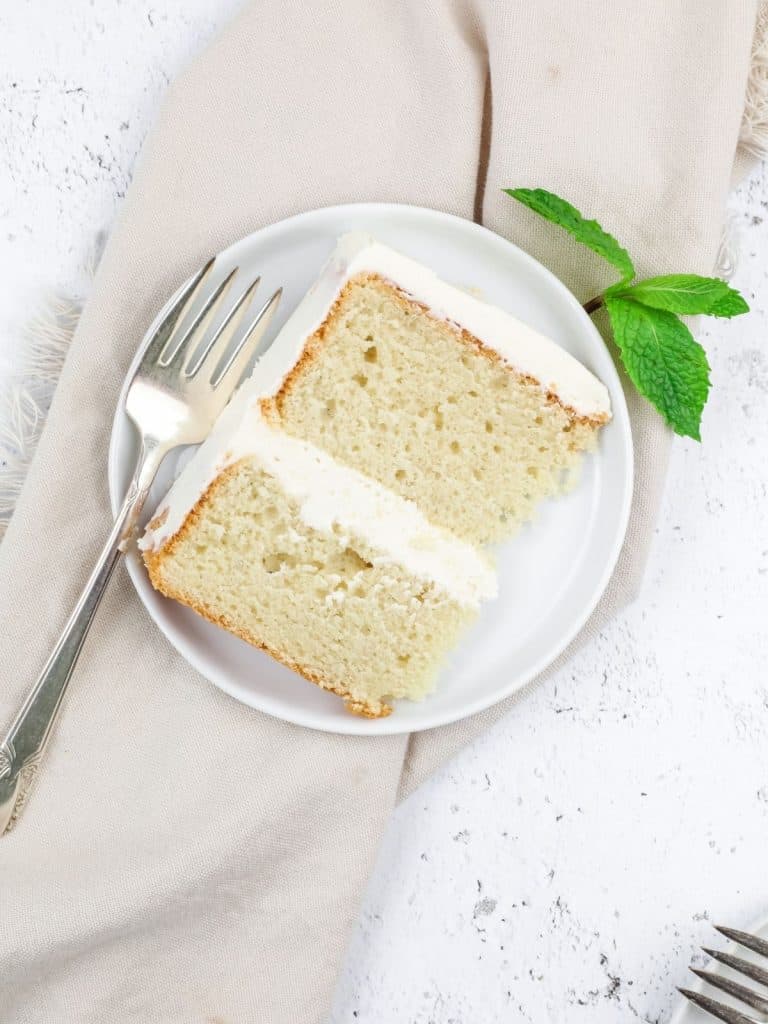 a perfect slice of gluten free vanilla cake with white buttercream frosting on a small round white plate with a fork and mint leaves