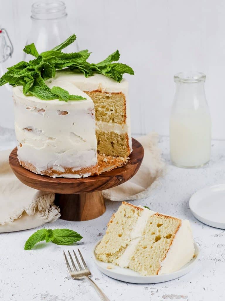 gluten free vanilla cake with white frosting on a wood cake stand with a slice cut out and resting on a small round white plate. mint leaves are covering the top of the cake