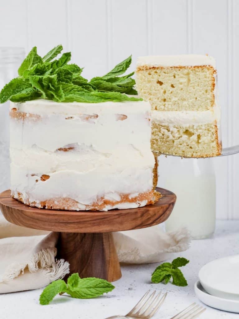 a slice of gluten free vanilla cake with white frosting is being removed from the cake and lifted with a sliver cake server. plates and forks and linen surround the cake on the kitchen countertop.