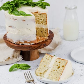 a gluten free vanilla cake rests on a wooden cutting board on a white surface covered in white frosting and mint leaves. a slice of cake rests on a round white plate with a fork beside it