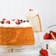 a gluten free angel food cake rests on a glass cake stand and is topped with whip cream and berries. a serving spoon is lifting a slice of cake away from the cake.
