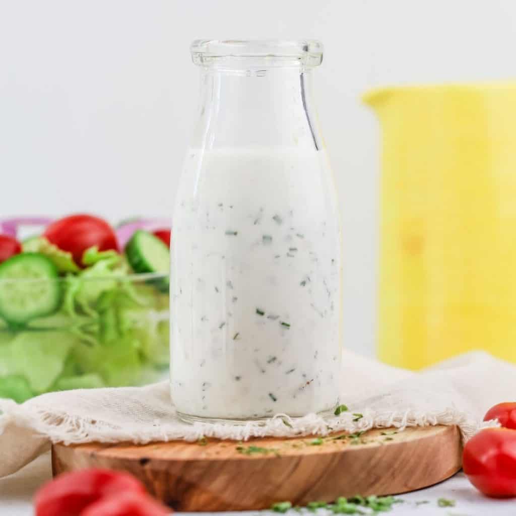 a small glass jar filled with vegan ranch dressing is sitting on a cutting board with fresh salad and tomatoes surrounding it