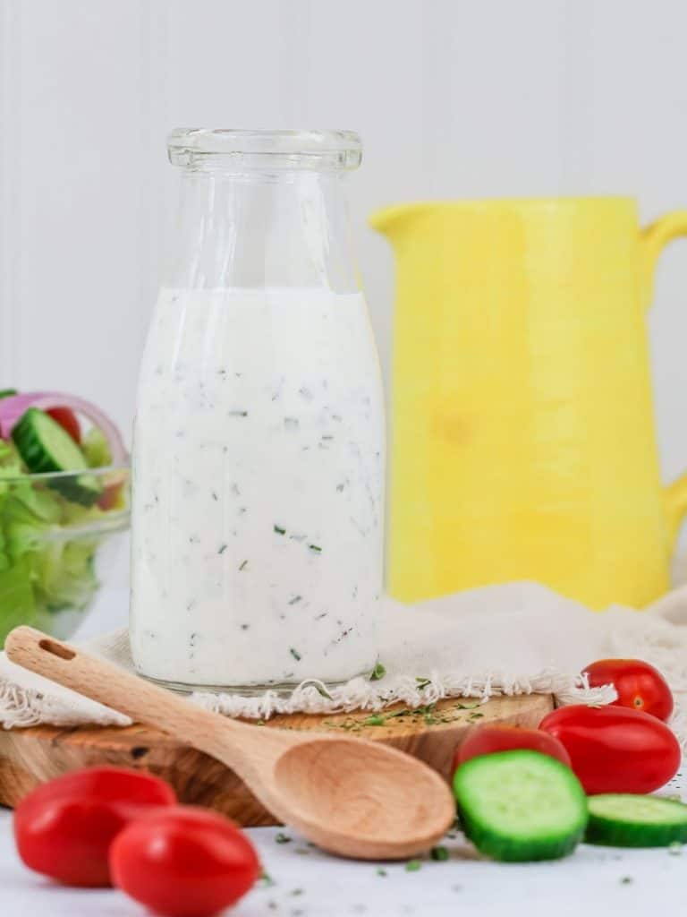 a glass bottle is filled with vegan ranch dressing that is resting on a wood cutting board. cucumbers and tomatoes surround the bottle. a small wood spoon and yellow pitcher are also seen.