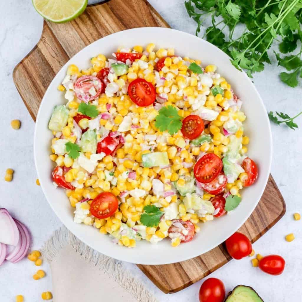 summer street corn salad is in a large round white bowl on top of a wood cutting board with different ingredients surrounding it like tomatoes, onion, and cilantro