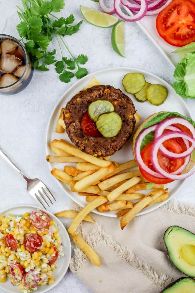 an open face black bean burger on a white plate surrounded by different toppings like tomatoes and pickles with a glass of soda and French fries