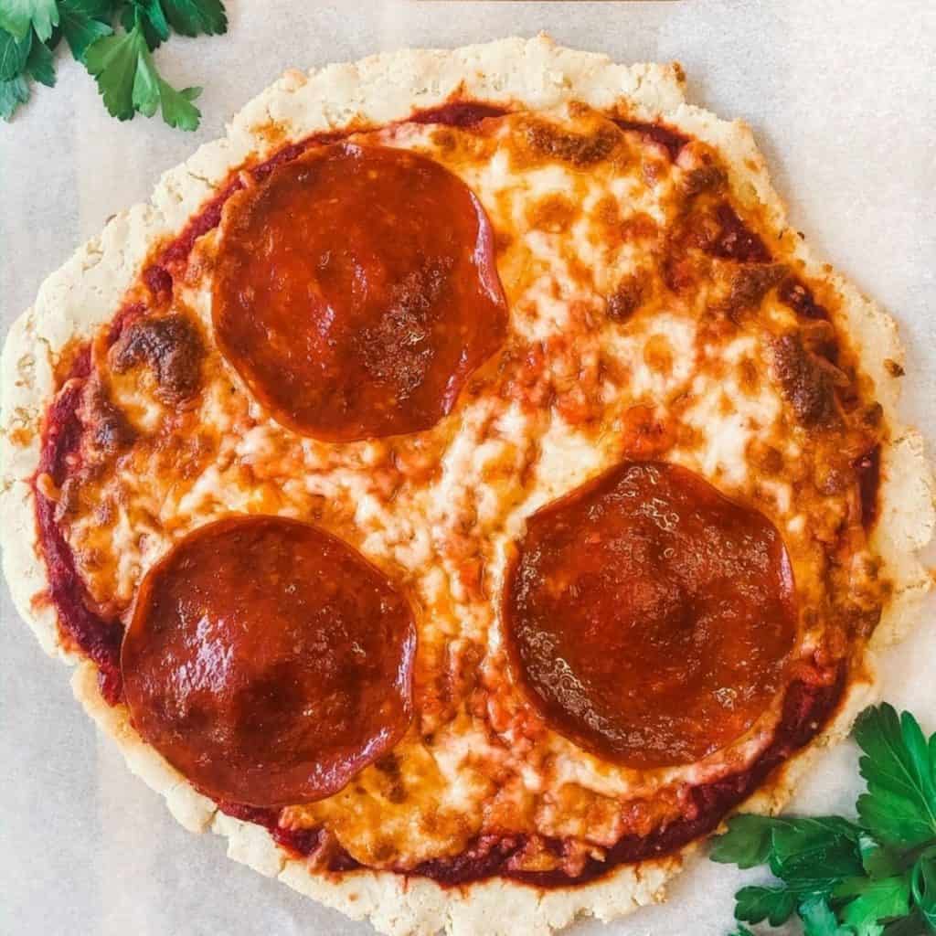 close up of a pizza covered in tomato free pizza sauce and large pepperoni on a white parchment paper with herbs surrounding it