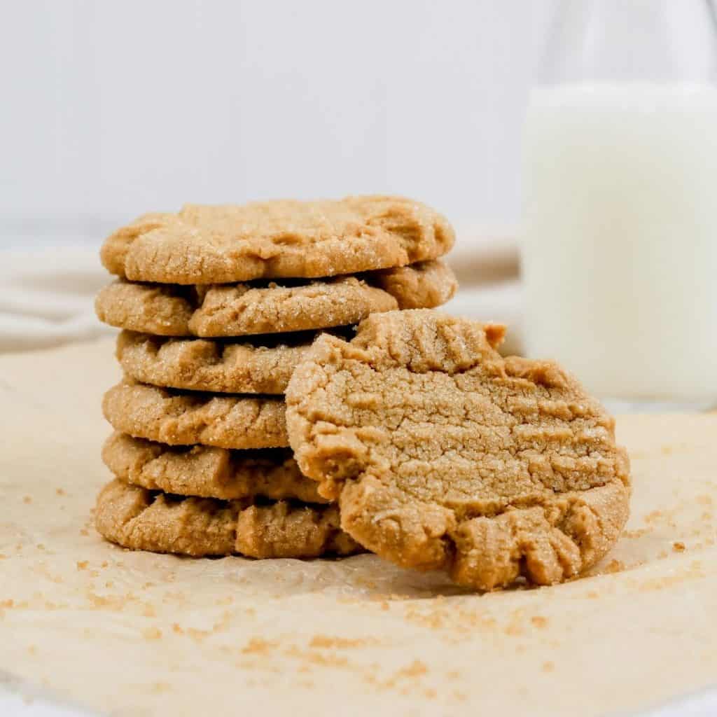 a stack of sun butter cookies is stacked with one leaning against them on parchment paper on the kitchen countertop with a glass of milk in the background