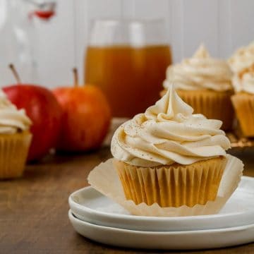 close up of apple cider cupcakes on a white plate in front of more cupcakes and apples