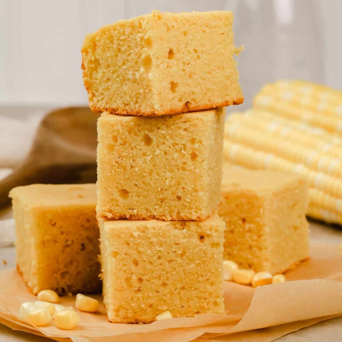 a stack of 3 slices of gluten free cornbread on parchment paper on the kitchen countertop surrounded by ears of corn
