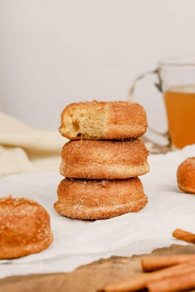 stack of 3 apple cinnamon donuts on white parchment paper with more donuts and apple cider in the background