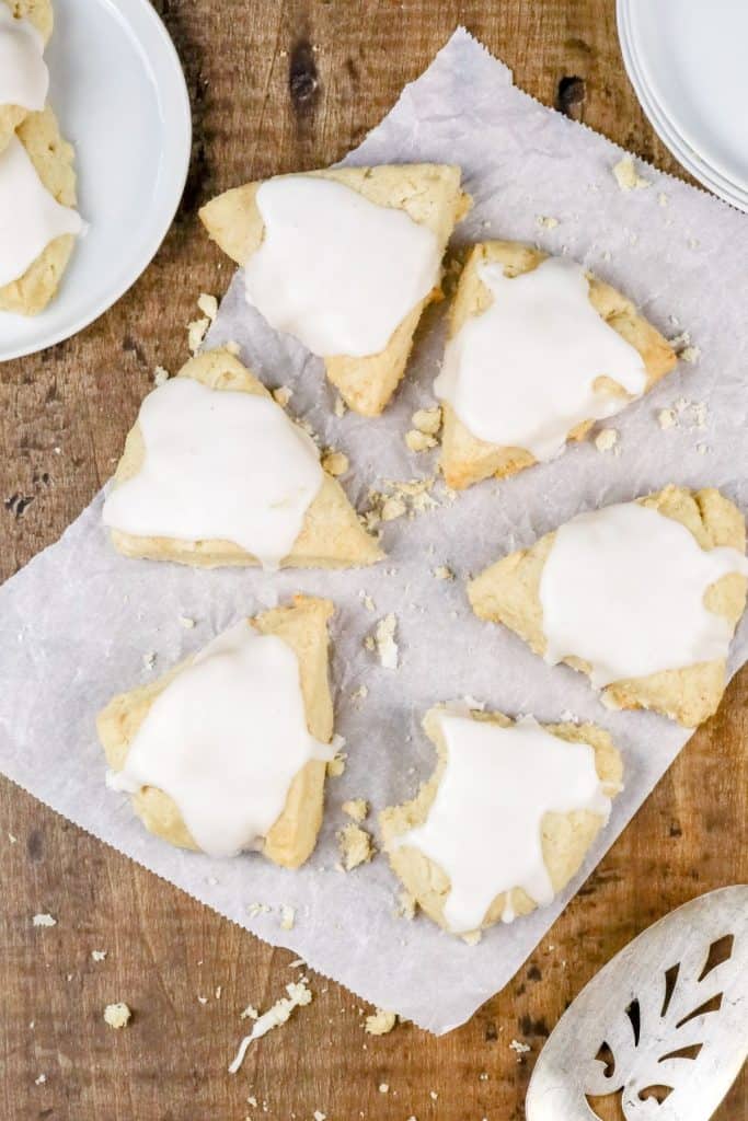 6 vanilla scones on a white parchment paper with plates, crumbs, and more scones surrounding them