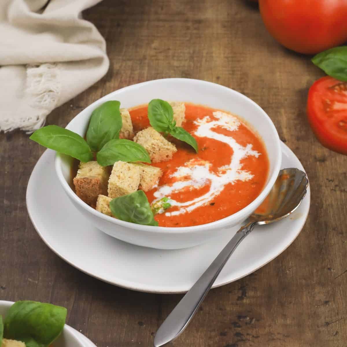 up close view of creamy vegan tomato soup in a white bowl on a white plate with basil leaves and a drizzle of cream on top