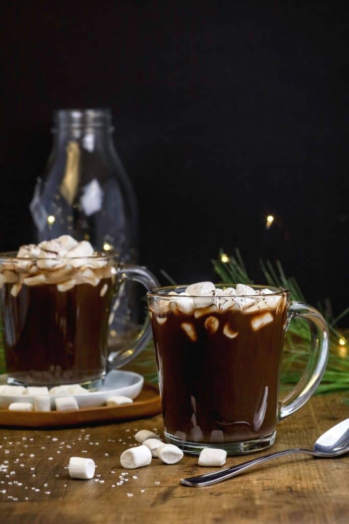 two glass mugs filled with hot chocolate and topped with marshmallows on a dark background