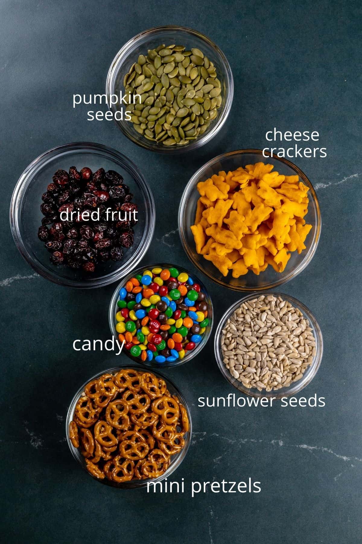 ingredients for nut free trail mix in various glass bowls on a dark countertop