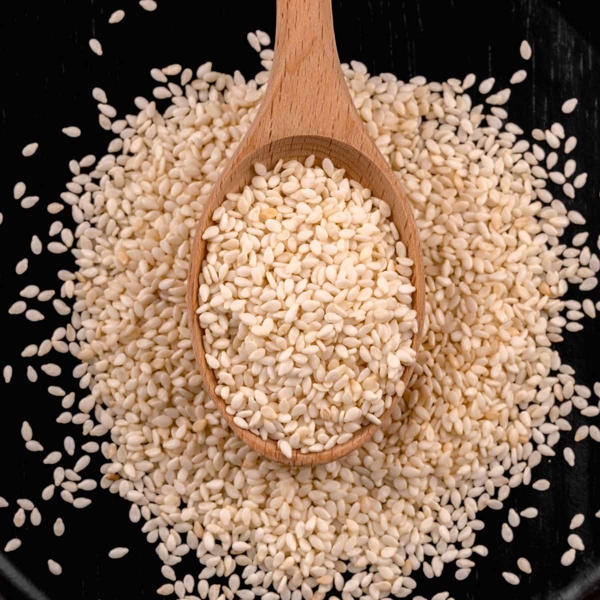 wood spoon full of sesame seeds on a black plate that is also filled with sesame seeds