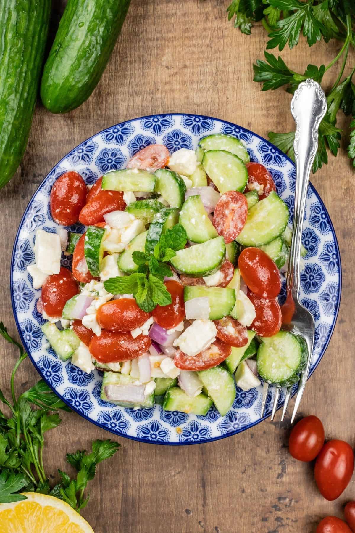 up close fresh cucumber salad in a blue and white bowl with ingredients surrounding it and a fork in the bowl