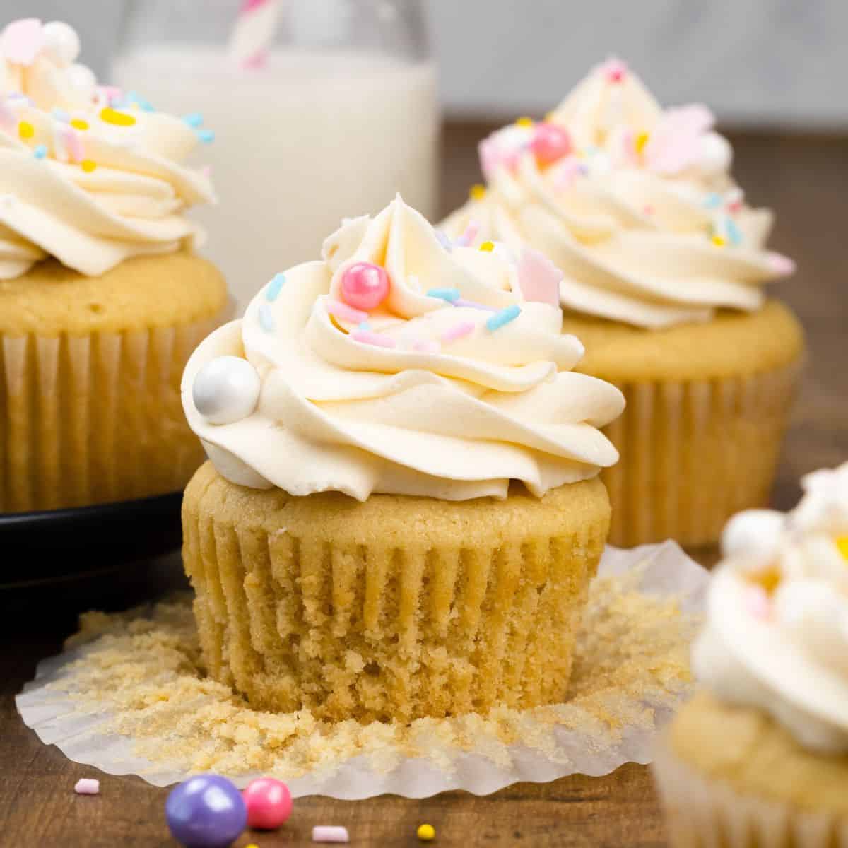 close up of an unwrapped vegan vanilla cupcake with vanilla frosting in a swirl on top and covered with sprinkles and more cupcakes surrounding it