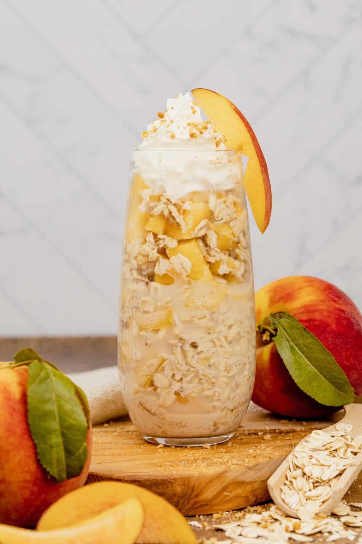 peach overnight oats in a glass cup with whip cream and a slice of peach on the side. More peaches surround the cup and a spoonful of oats can be seen.