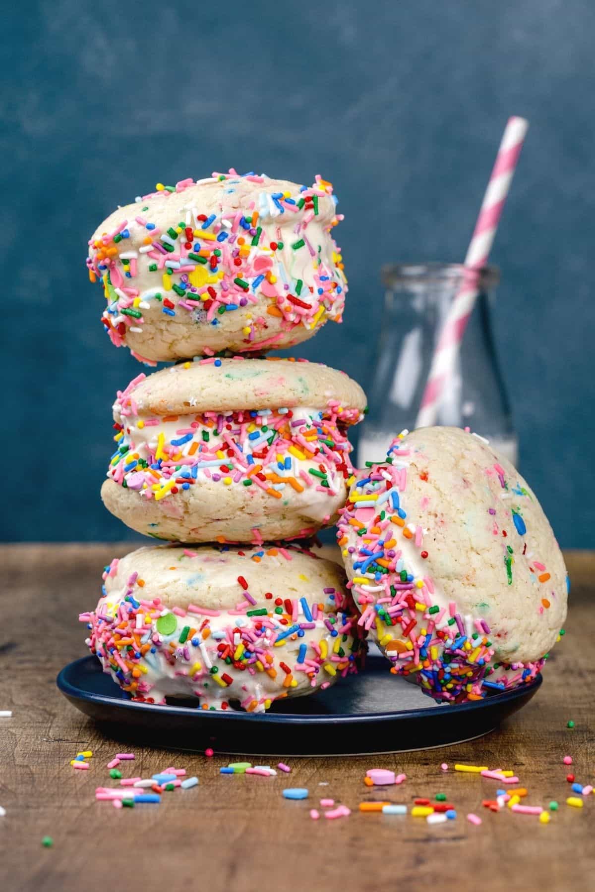 a stack of 4 ice cream sandwiches with rainbow sprinkles on a dark blue plate in front of a glass of milk with a pink straw blurred in the background