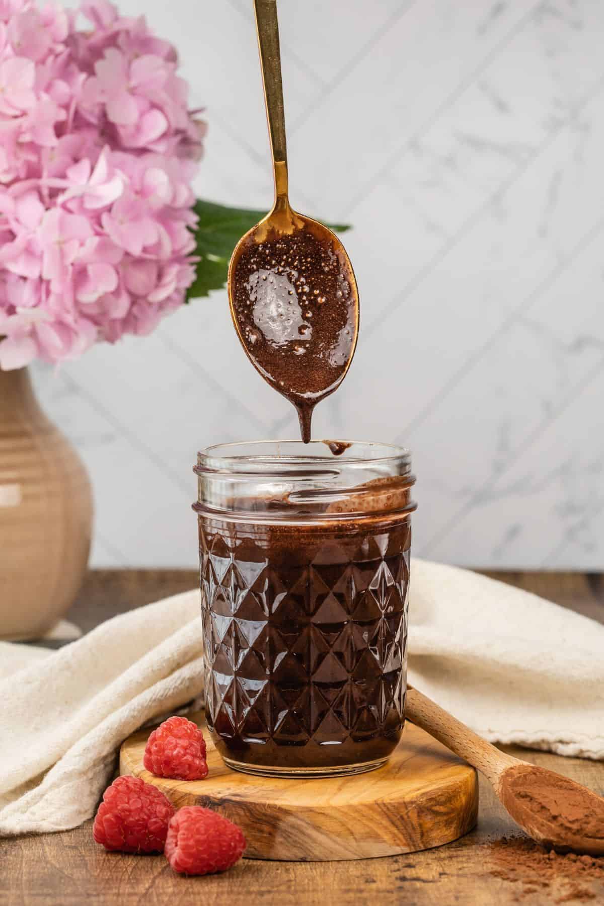 mason jar filled with vegan chocolate sauce with a spoon pouring more into the jar. a vase of flowers is blurred in the background and raspberries are blurred in the foreground
