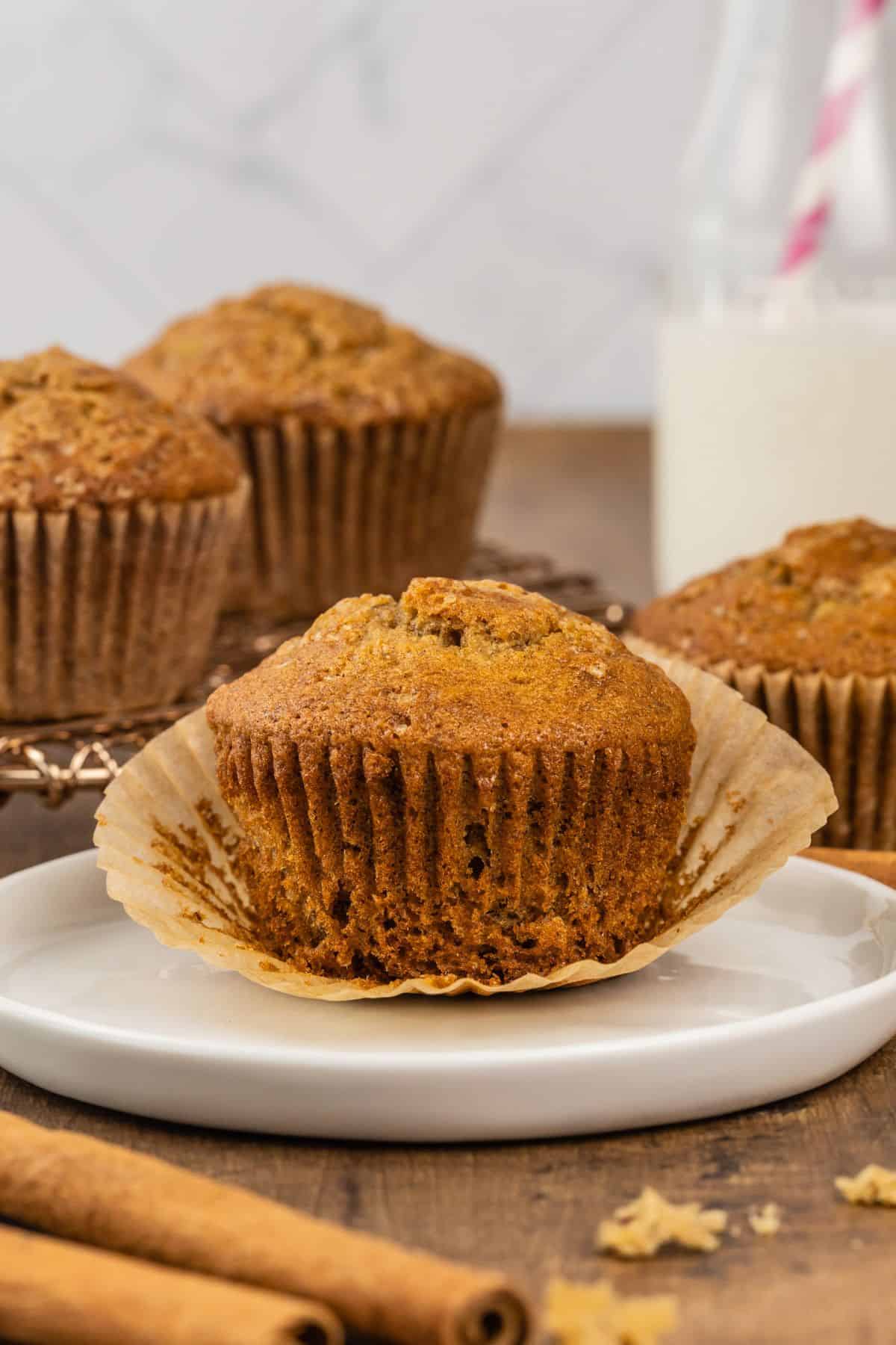 a single unwrapped gluten free banana muffin rests on a round white plate with more muffins and cinnamon sticks blurred in the background and foreground