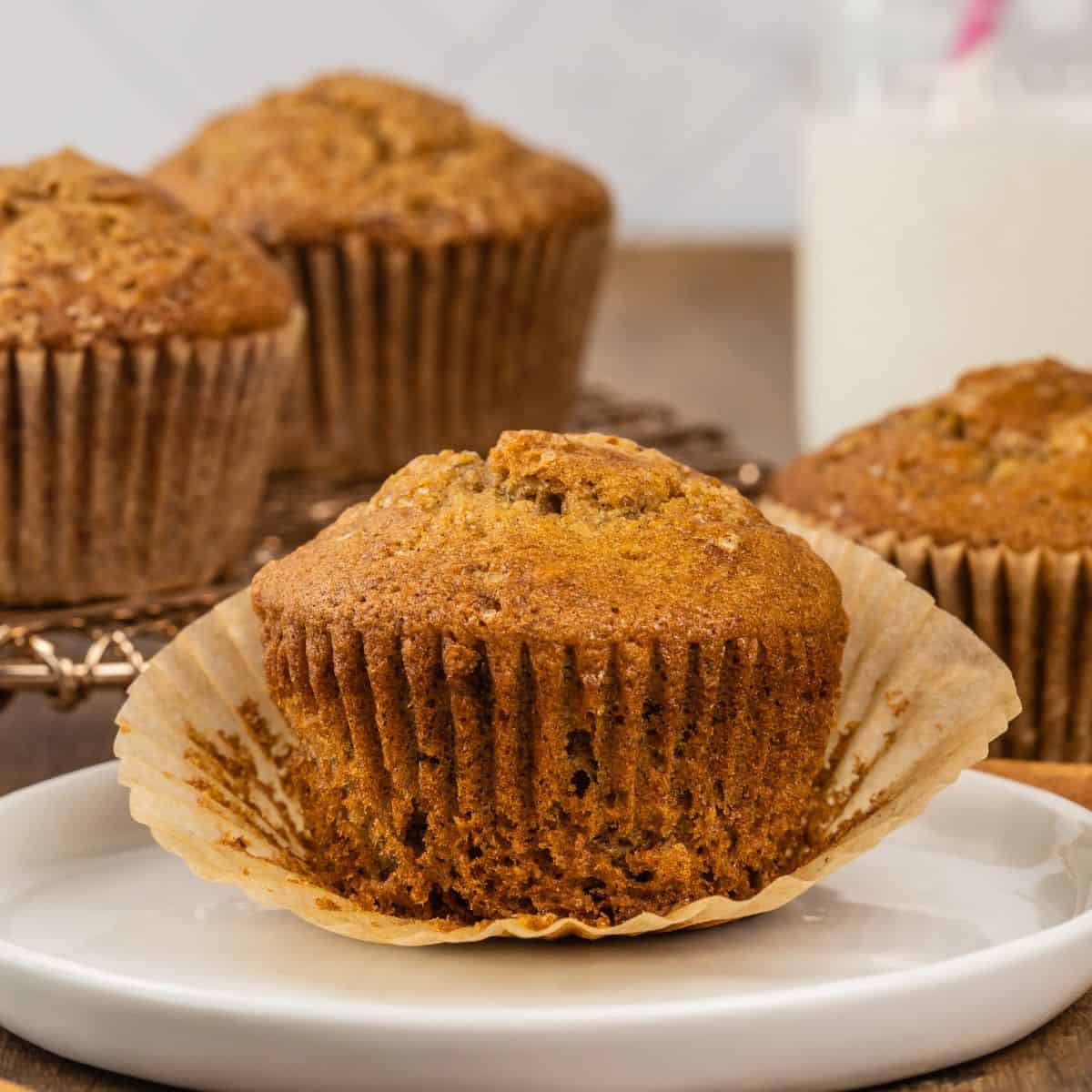 an unwrapped gluten free banana muffin sits on a white round plate with more muffins blurred in the background