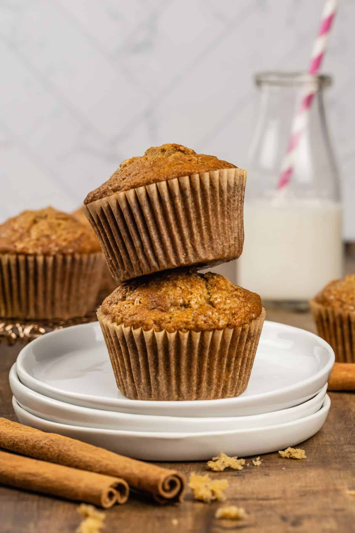 a stack of two gluten free banana muffins on top of a stack of small round white plates on the kitchen table with more muffins and a glass of milk with a pink straw blurred in the background