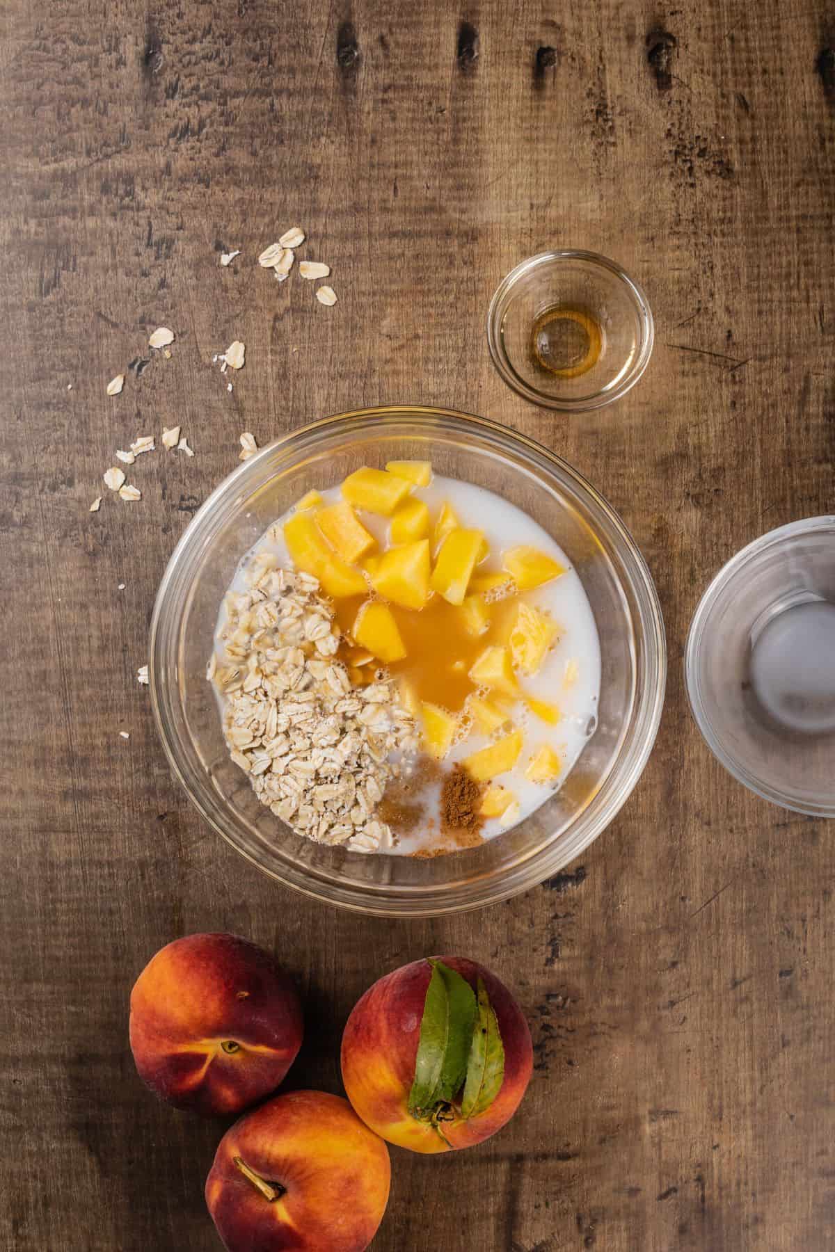 a glass bowl filled with all the ingredients for peach overnight oats on a wood tabletop